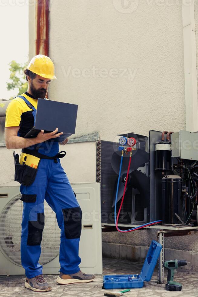 Certified expert commissioned for outside air conditioner routine check up, imputing data on laptop. Trained mechanic doing hvac system safety check, looking for defective internal parts photo