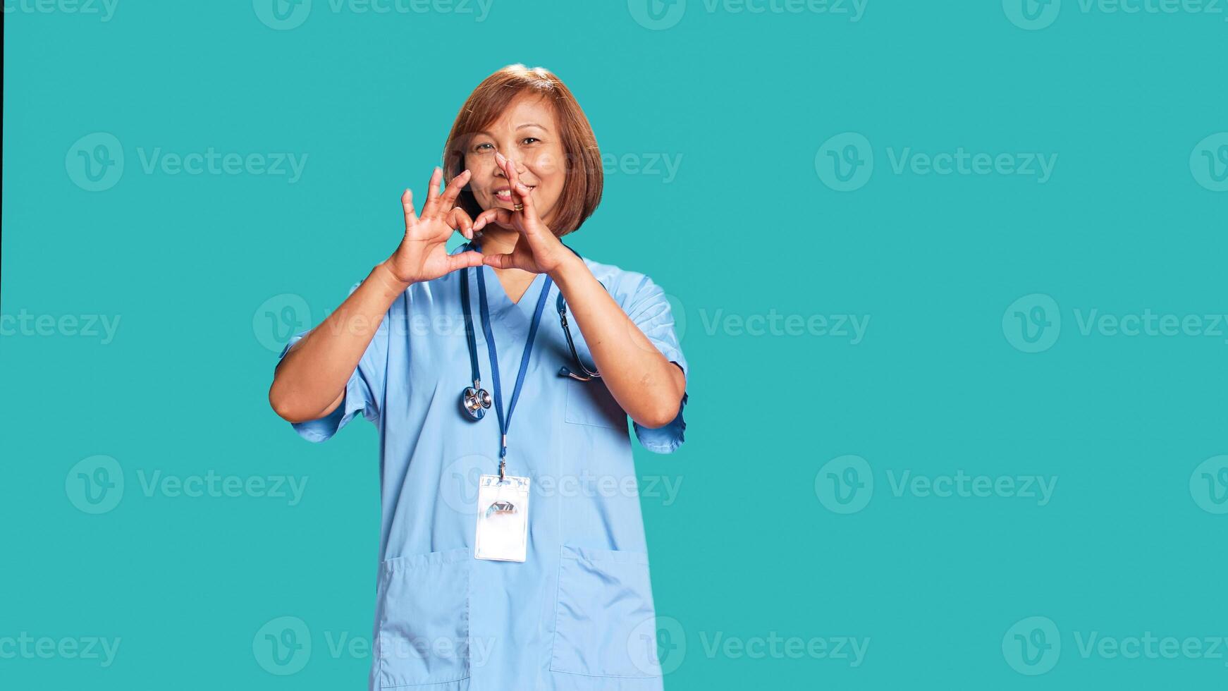 Portrait of cheerful healthcare professional doing heart symbol shape with hands. Licensed asian nurse showing love gesturing while at work, isolated over blue studio background photo