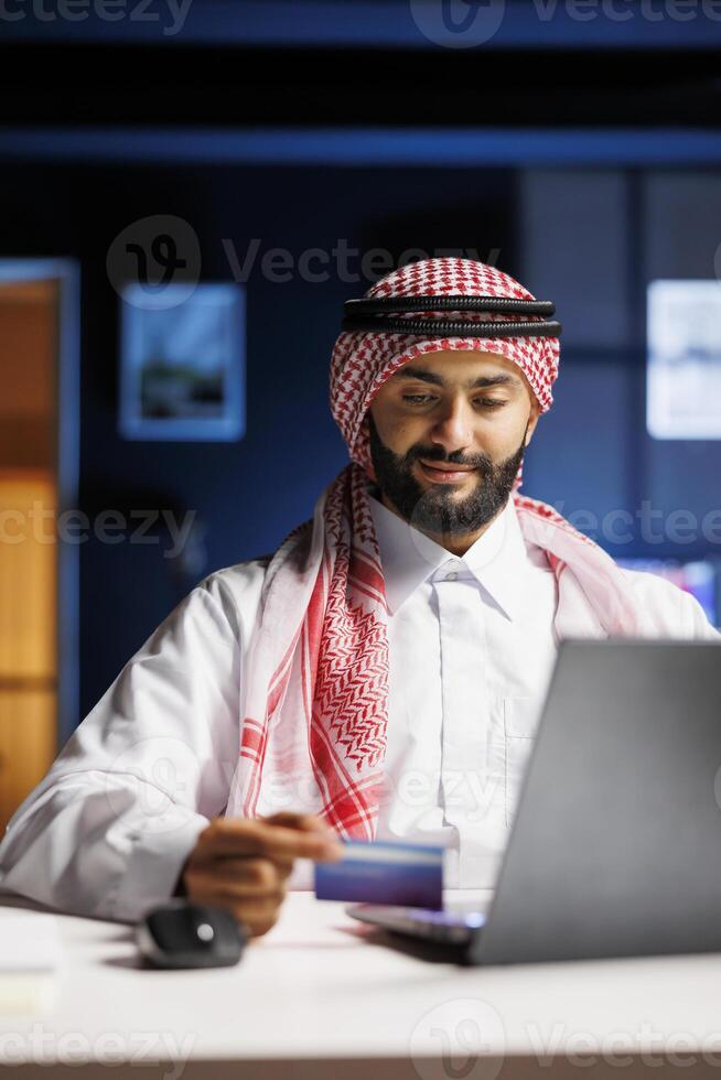 A focused Arabian businessman in Islamic attire sits at a modern desk, typing on a laptop. He efficiently works, utilizing wireless technology for online communication, surfing the net, and conducting online shopping and payments. photo