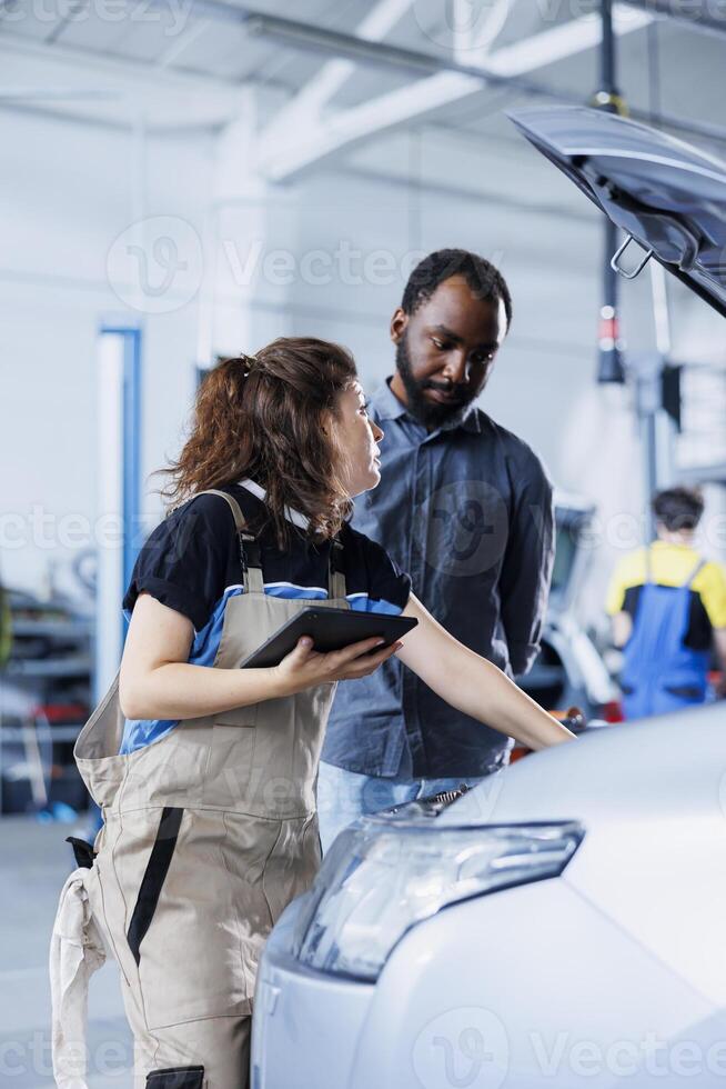 Mechanic in car service ordering new ignition system for damaged vehicle using tablet. Employee next to BIPOC client looking online for components to replace old ones in malfunctioning automobile photo