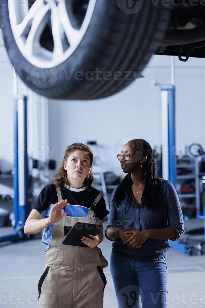 Mechanic at auto repair shop conducts annual vehicle checkup, informing client about needed wheels replacement. Garage worker talks with customer underneath car after finishing inspection photo