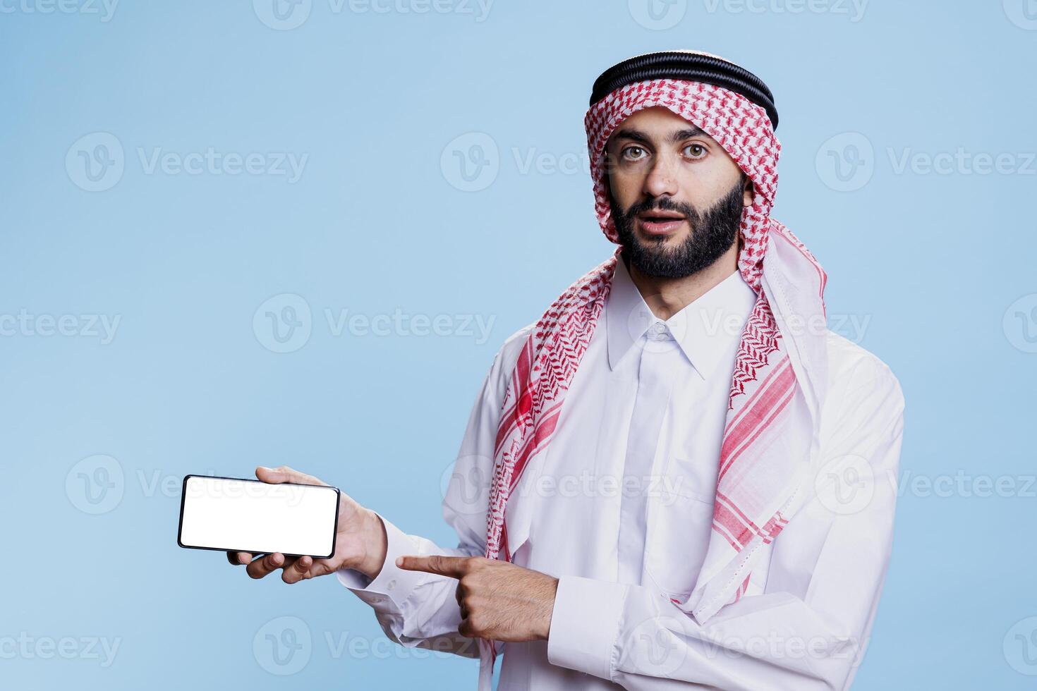 musulmán hombre participación teléfono inteligente con blanco pantalla en horizontal modo y señalando con dedo. árabe persona en tradicional ropa demostración vacío pantalla táctil y mirando a cámara foto