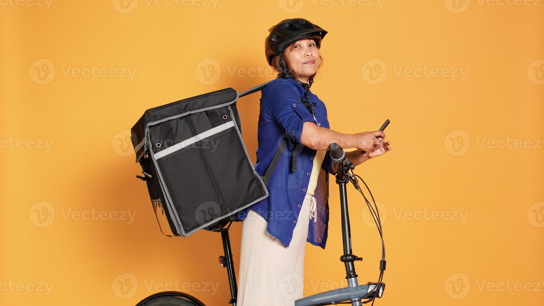 BIPOC courier woman taking break between takeout meal deliveries, texting friends. Cyclist waiting to receive next customer food order, isolated over orange studio background photo