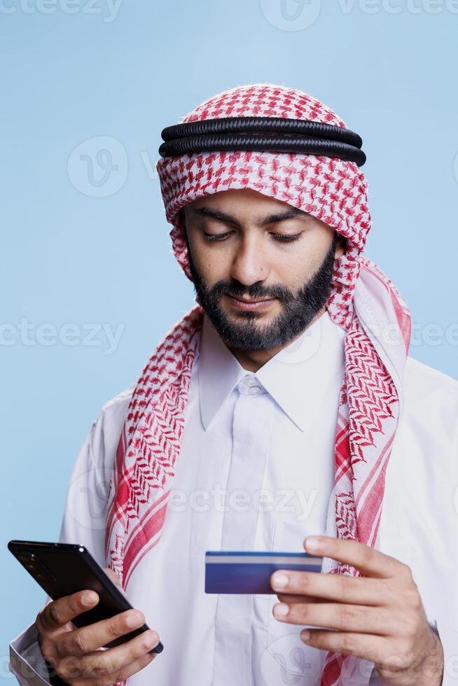 Man in traditional muslim clothes looking at credit card while making electronic payment on smartphone online store. Arab wearing ghutra and thobe processing purchase in internet shop photo