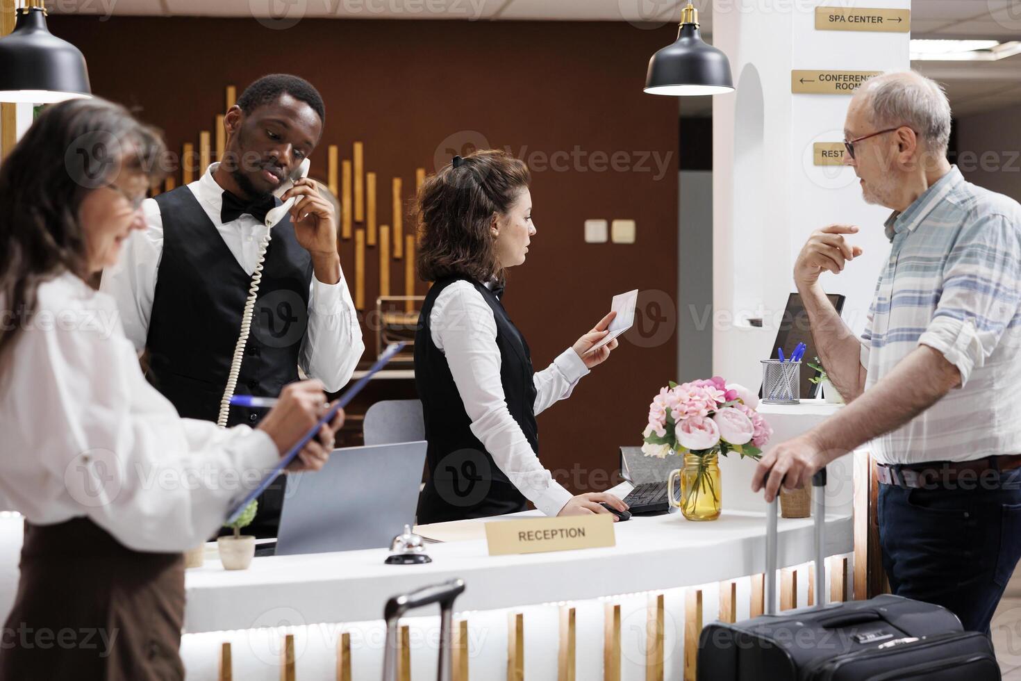 While african american staff answers phone, female concierge verifies id of elderly male traveler at hotel front desk. Retired senior woman fills out form as old man waits for his passport at counter. photo