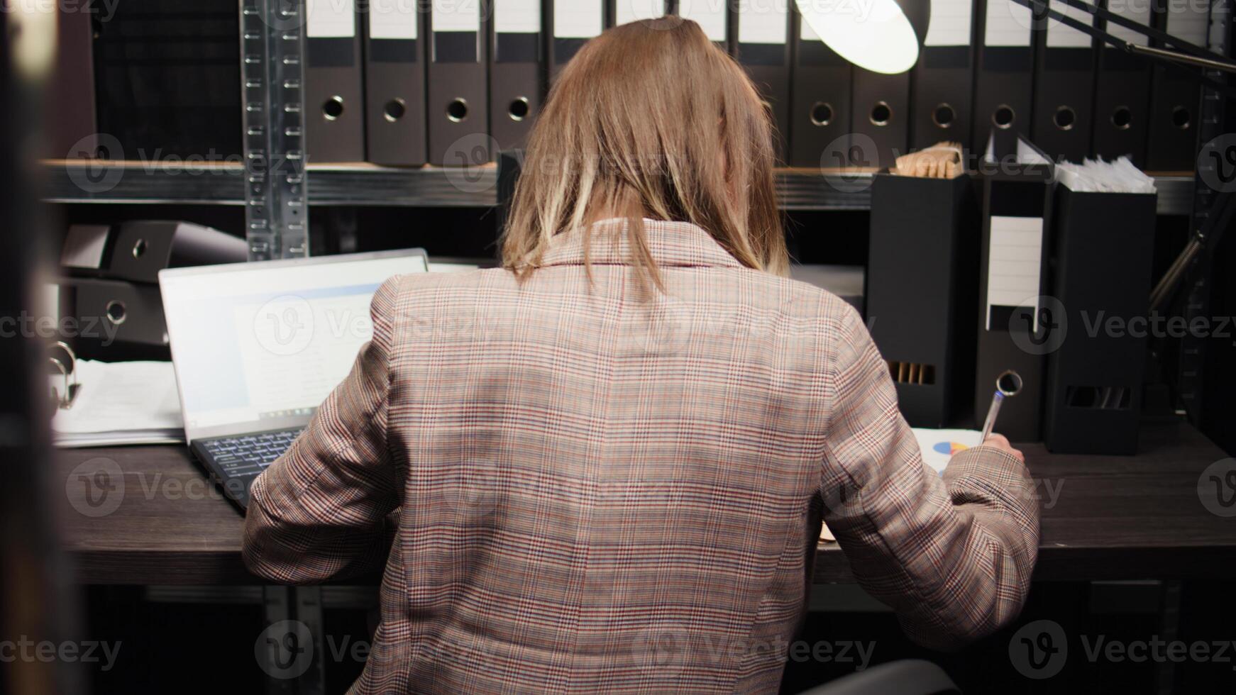 Female crime expert examining papers to put the pieces together and analyzing forensic records while taking notes. Caucasian private detective looking into a case of organised crime. Back-view shot. photo