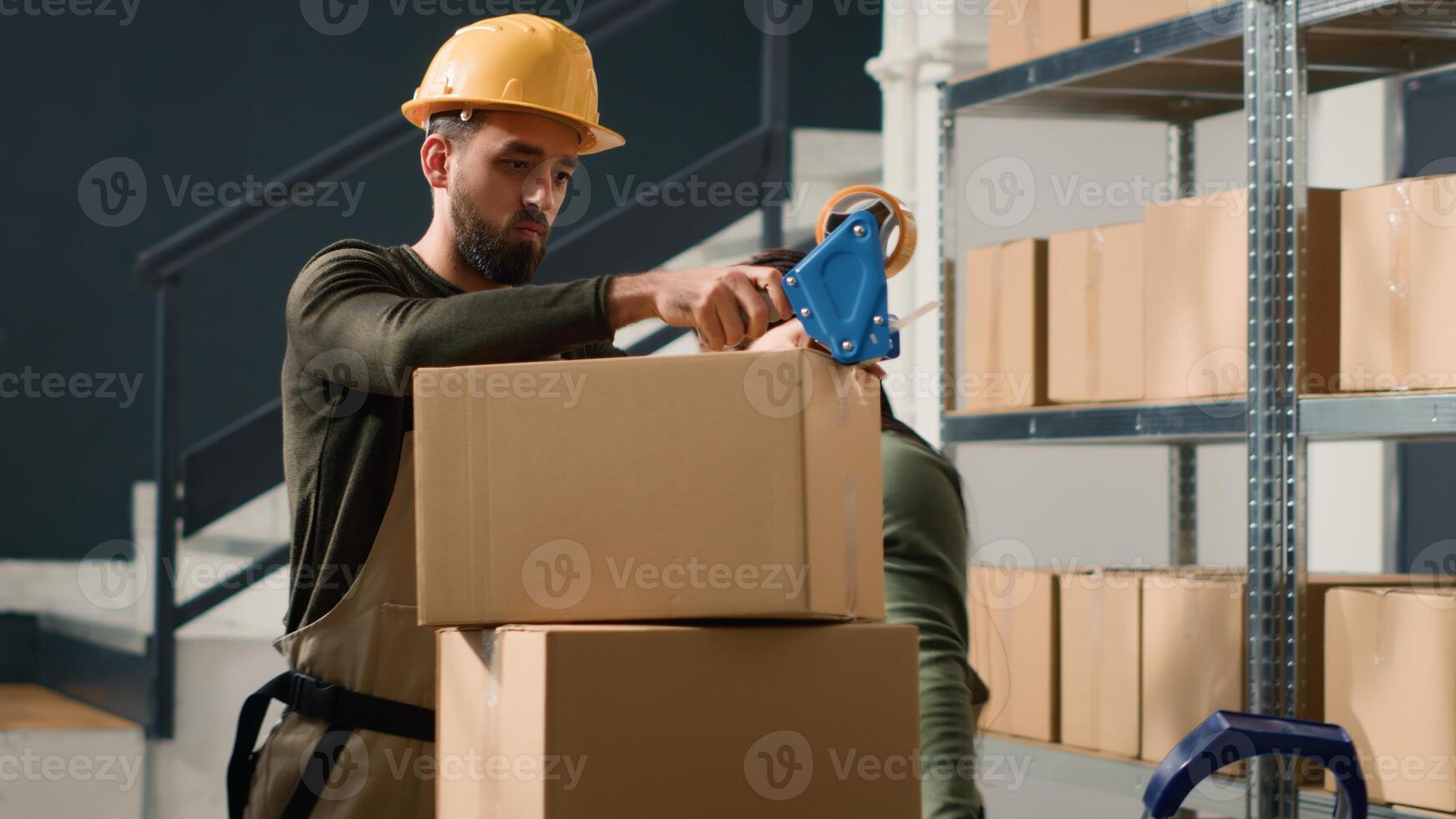 Manager wearing protective gear in warehouse teaching african american trainee how to seal cardboard box packages. Storage site personnel properly securing goods to avoid issues during shipping photo