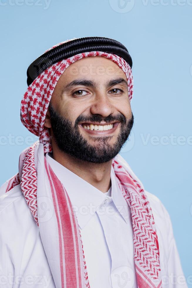 Smiling arab man wearing traditional headscarf looking at camera with cheerful face expression. Joyful muslim person in ghutra headdress posing for studio portrait on blue background photo