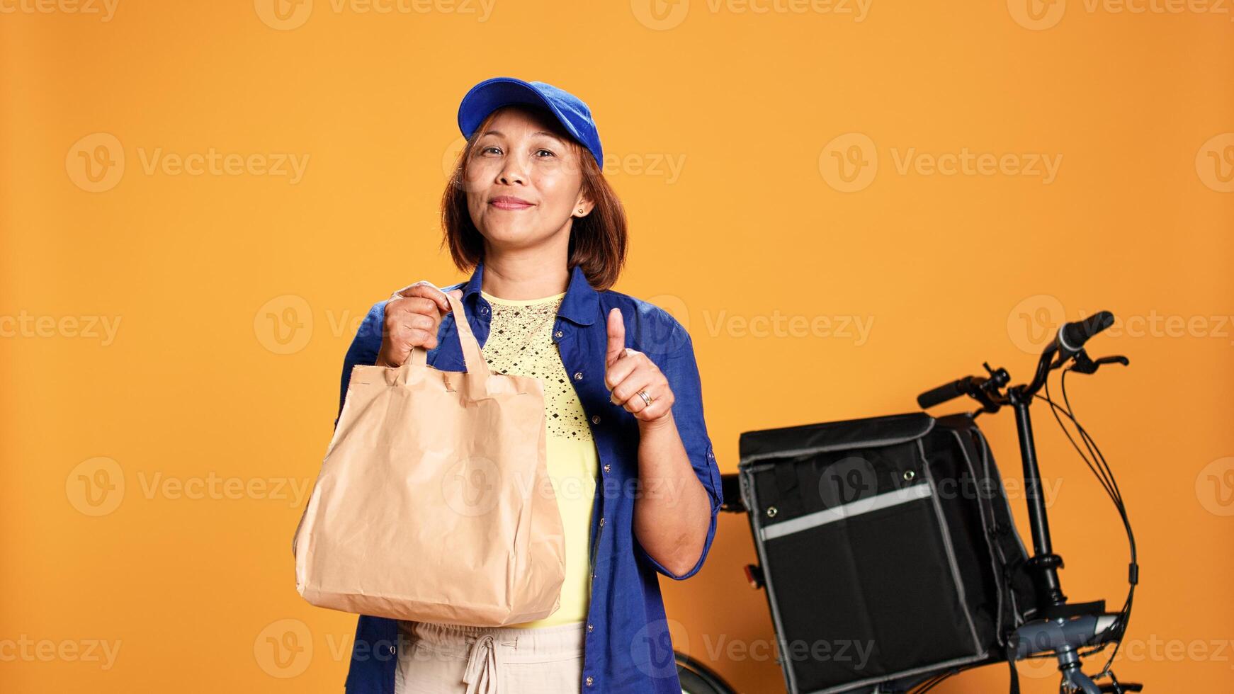 Slow motion portrait shot of friendly courier waiting for customer to answer door, showing thumbs up sign. Food delivery cyclist handing lunch bag to customer, isolated over studio background photo