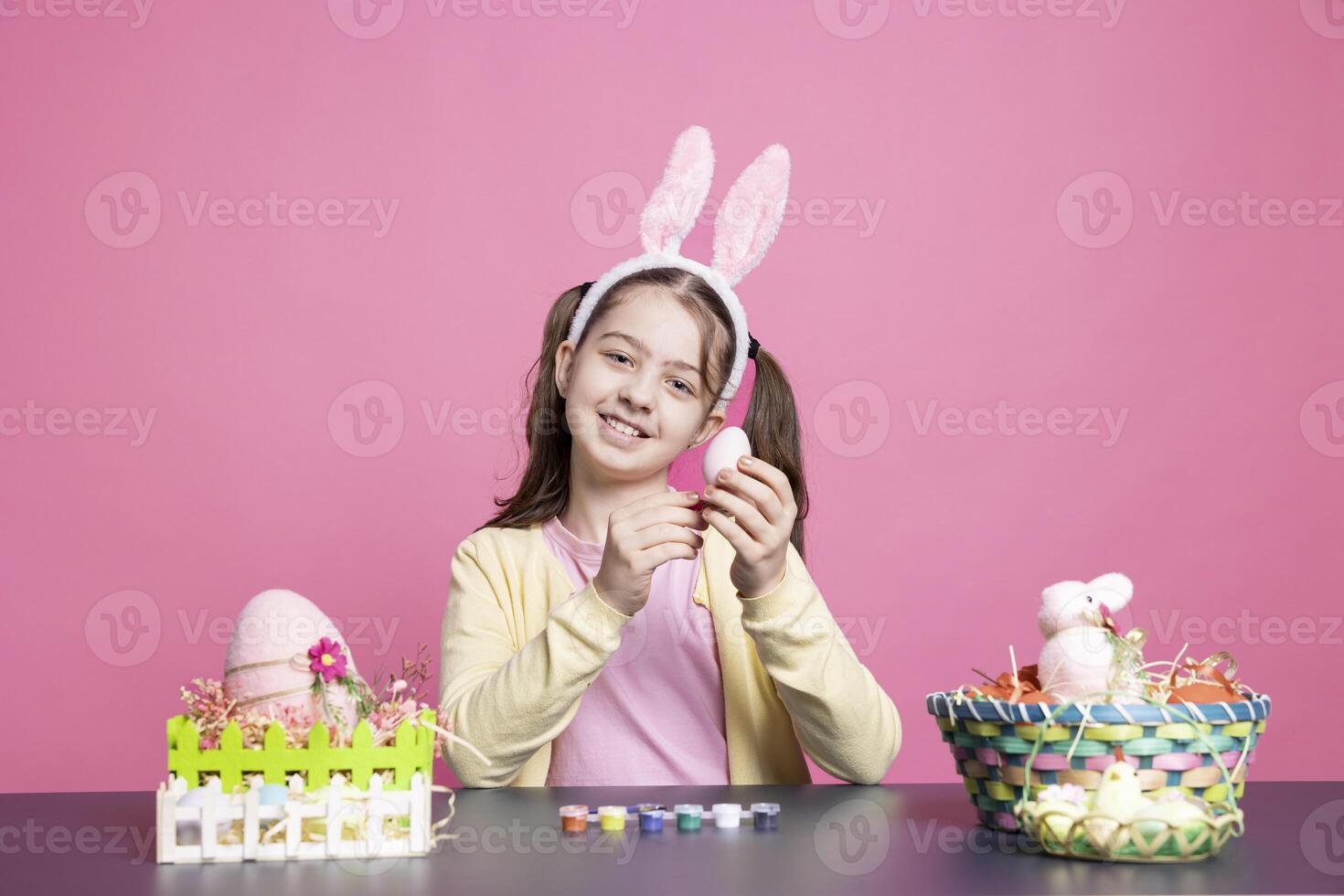 Delighted youngster wearing bunny ears decorating eggs at workspace, making handcrafted decorations in celebration of Easter holiday. Adorable kid having fun with paintbrush to decorate ornaments. photo
