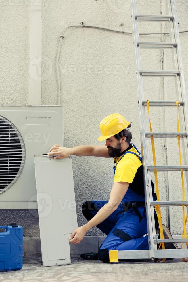 Experienced serviceman starting work on broken air conditioner, using pneumatic drill to pull apart condenser metal coil panel. Certified electrician taking apart hvac system to check for leaks photo