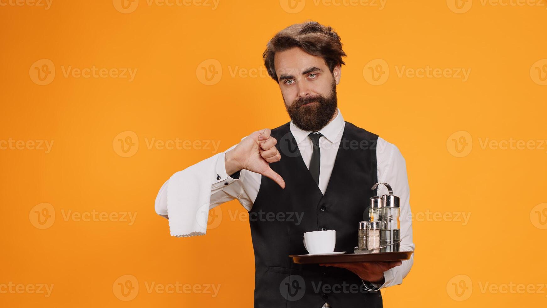 Restaurant employee shows dislike symbol on camera, giving thumbs down against yellow background. Classy fine dining butler presents disapproval and acting displeased, holds platter with drinks. photo