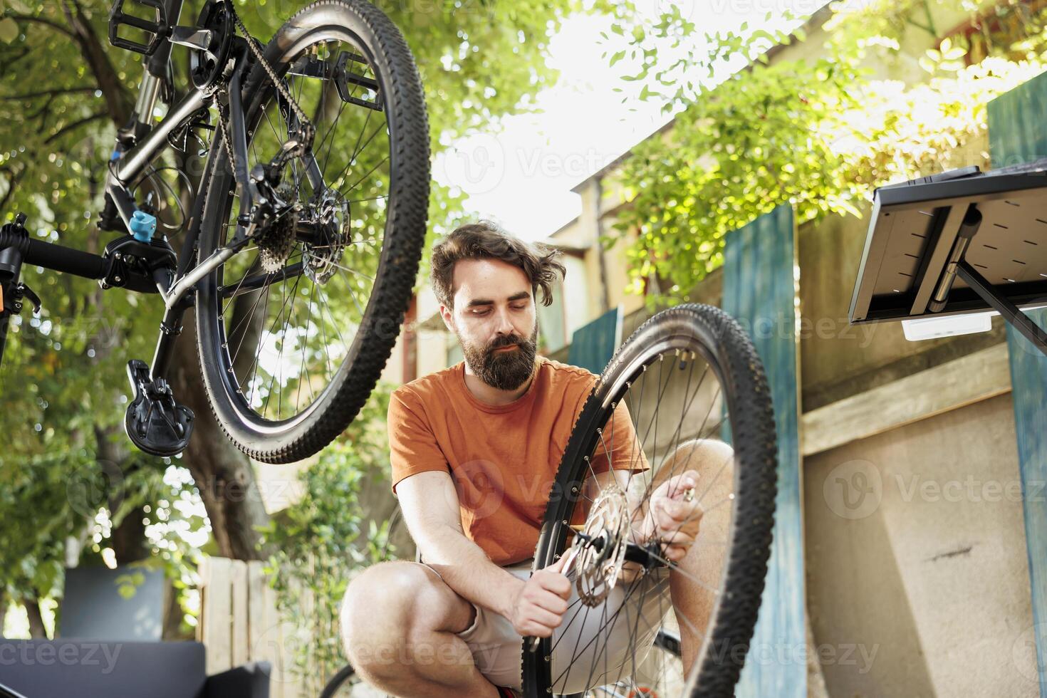 Sporty and dedicated man adjusting bicycle gears with professional equipment in home yard. Active caucasian male repairs and maintains bike components outside using specialized tools. photo