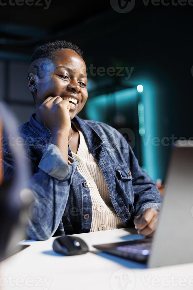 Close-up of black woman laughing while using a digital laptop at the table. African american lady with hand on her face, joyfully utilizing modern technology, browsing through her personal computer. photo