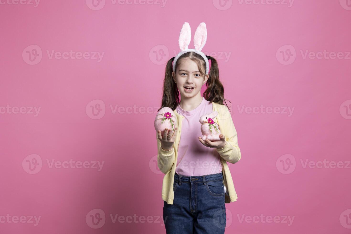 pequeño niña con conejito orejas participación rosado Pascua de Resurrección decoraciones en frente de cámara, presentación su hecho a mano huevo y relleno Conejo. joven niño sonriente en estudio y celebrando primavera festividad. foto