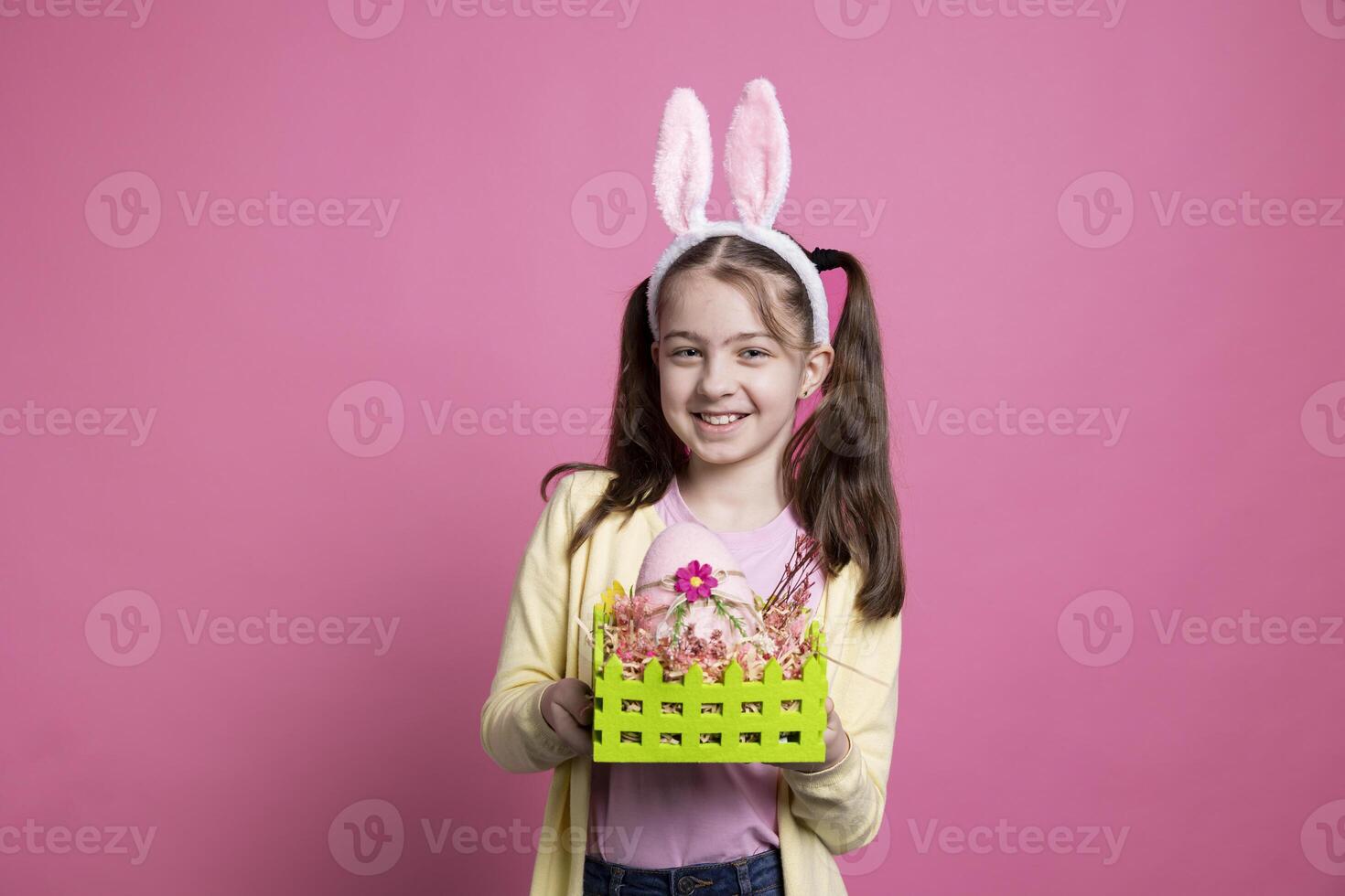 Positive playful kid wearing bunny ears and pigtails in studio, posing with festive decorated eggs in a colorful basket. Young cheerful child happy about easter decorations and festivity. photo