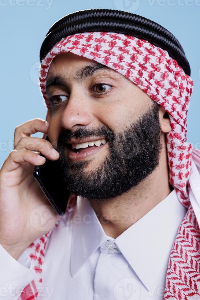 Excited muslim man wearing traditional checkered headscarf answering smartphone call with smiling expression. Cheerful arab dressed in islamic headdress speaking on mobile phone closeup photo