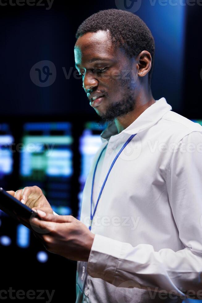 African american admin using laptop to review energy consumption across server rack parts. Professional making sure data center electronics temperature sensors are in perfect working condition photo