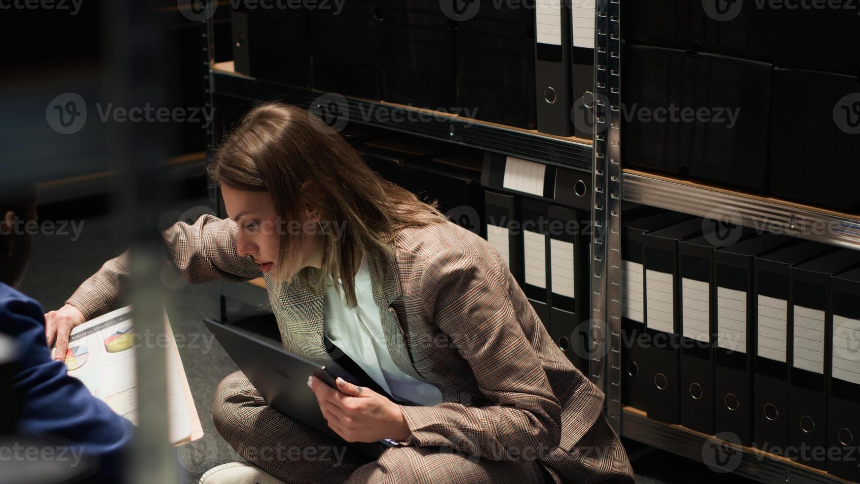 Policewoman conducts an investigation while seated on floor of archive room with papers, heaps of evidence, and documents in folders. Female detective gathering case information with laptop. photo