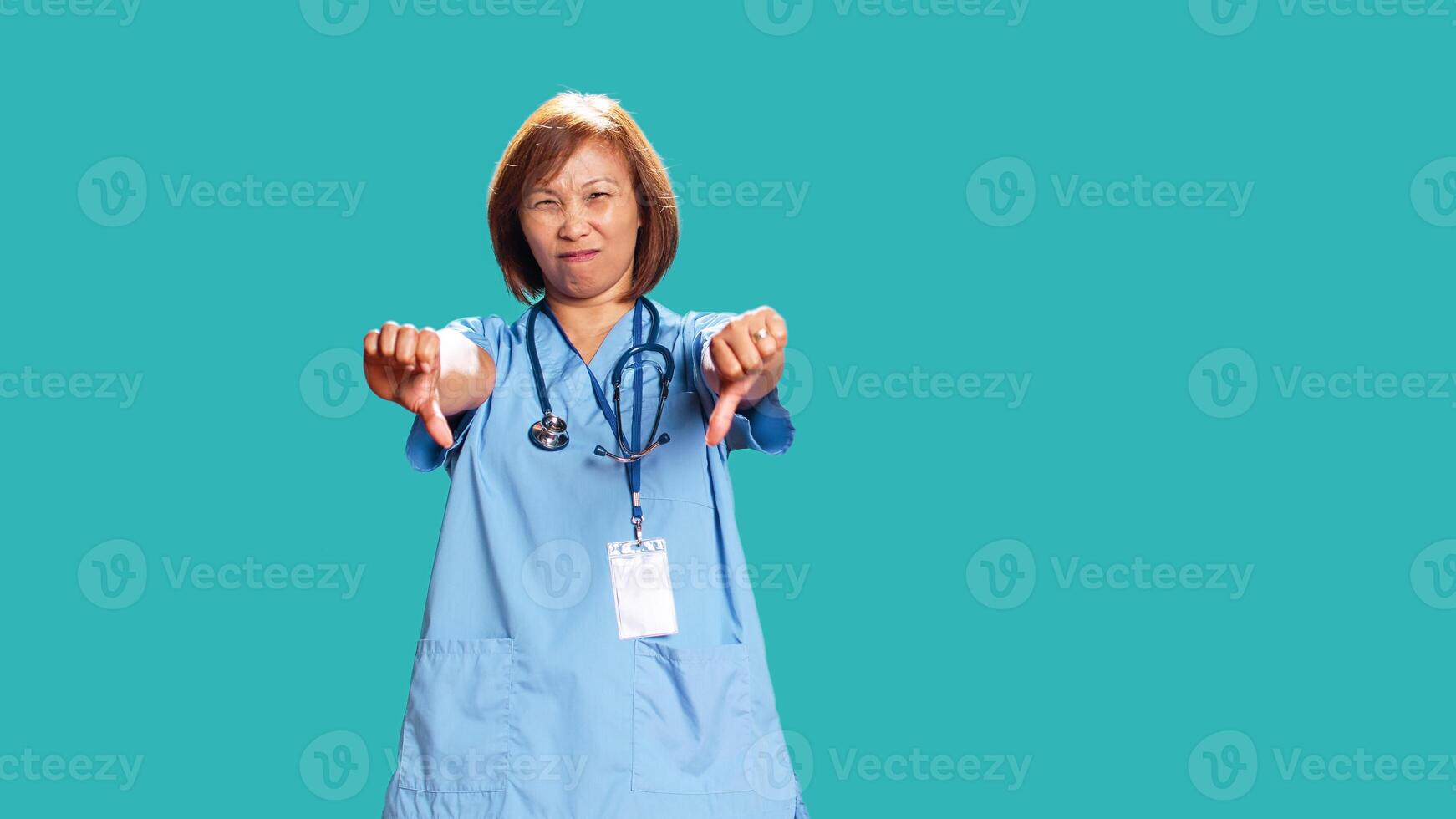 Upset asian nurse showing thumbs down signs while at work, isolated over blue studio background. Dissapointed BIPOC healthcare professional doing frenetic disapproval gestures photo