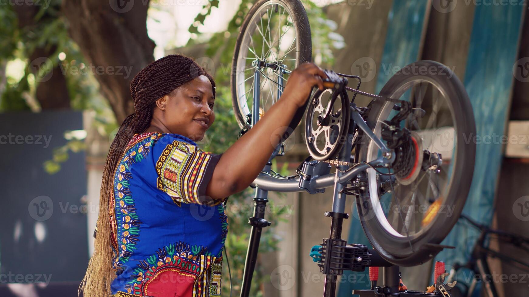 africano americano mujer atentamente comprobación bicicleta componentes asegurando suave y eficiente verano ocio ciclismo. sonriente hembra ciclista ejecutando bicicleta mantenimiento. vista lateral retrato disparo. foto