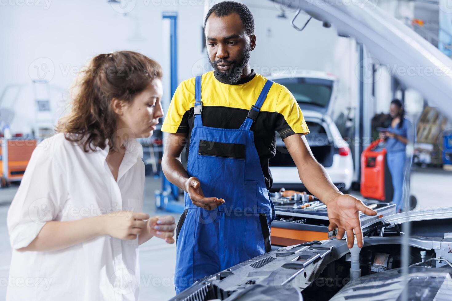 Repairman at auto repair shop conducts routine vehicle checkup, informing customer about needed engine fix. BIPOC garage worker chatting with customer after finishing car maintenance photo