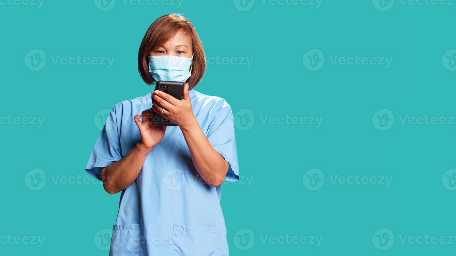 Happy relaxed nurse taking break between hospital shifts, texting friends, having fun online. Smiling woman wearing protective face mask using gadget isolated over blue studio background photo