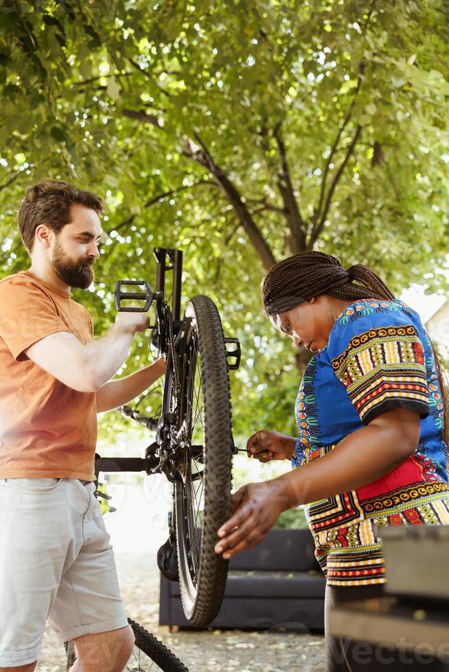 Sporty multiethnic couple inspecting and repairing bike tire and chain in home yard. Healthy and active two persons working outdoors on bicycle maintenance with expert work tool. photo
