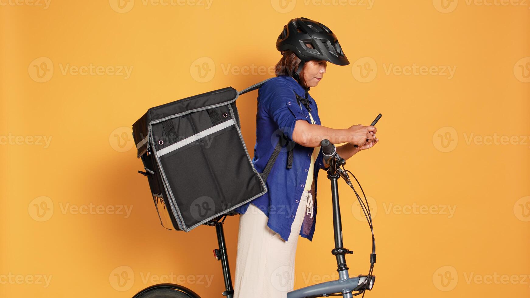 Asian cyclist delivering takeaway food to customers, imputing next location data in phone app. Courier woman riding bike, wearing safety helmet and thermal backpack, isolated over studio background photo