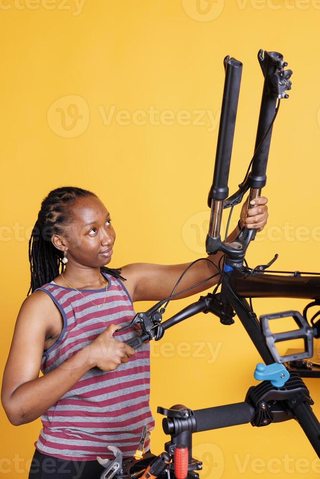 Determined african american lady adjusting and securing bicycle headset and handlebar in front of yellow background. Disassembled bike being repaired by sports-loving female for leisure cycling. photo