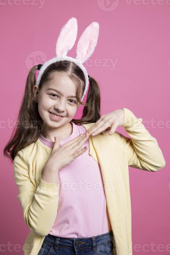 Cute young girl dancing and fooling around on camera, wearing bunny ears and pigtails over pink background. Carefree schoolgirl feeling joyful and excited about easter celebration in studio. photo