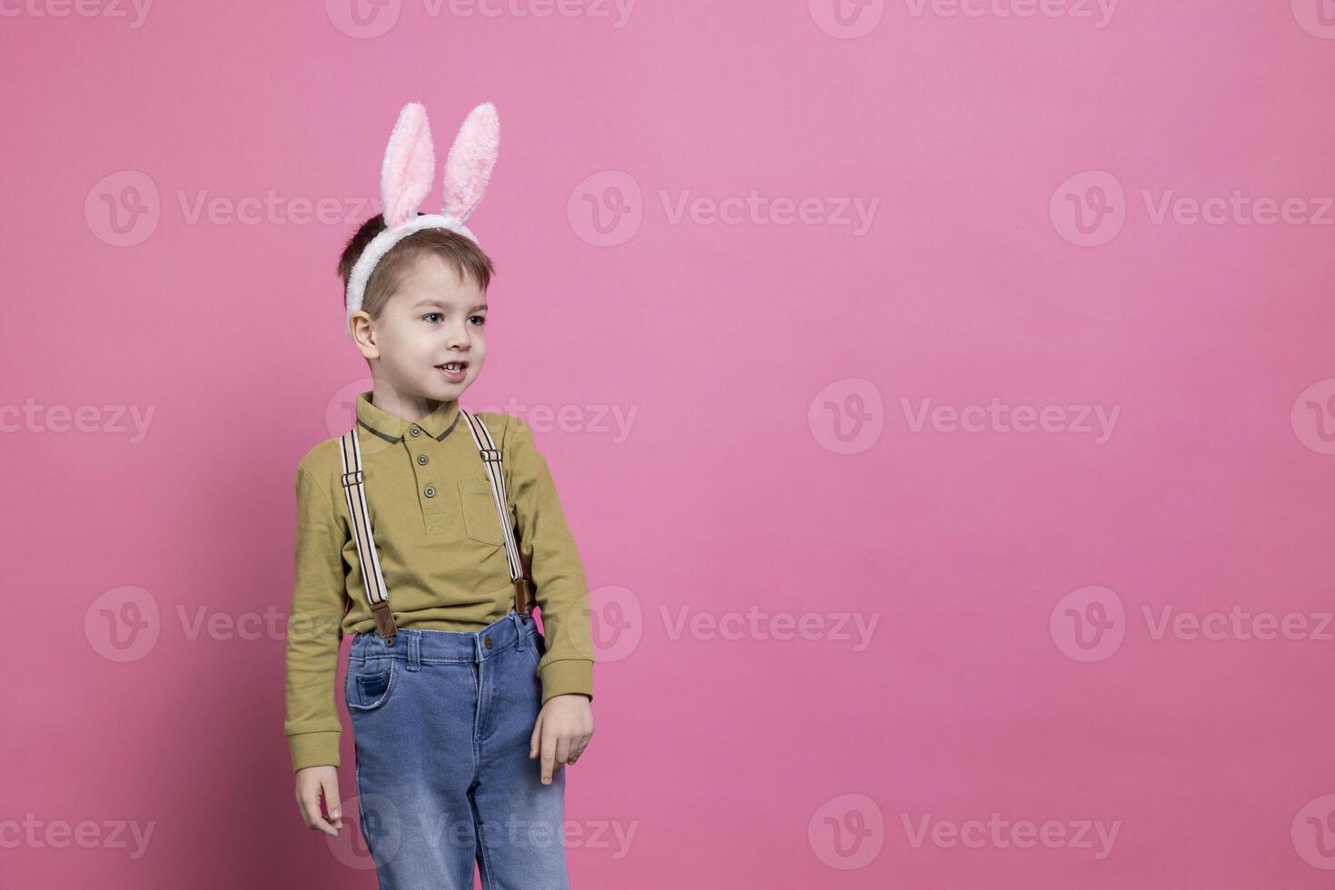 Little boy being cute with bunny ears in front of the camera, feeling excited about easter celebration event and receiving presents. Cheerful small kid feeling joyful against pink background. photo