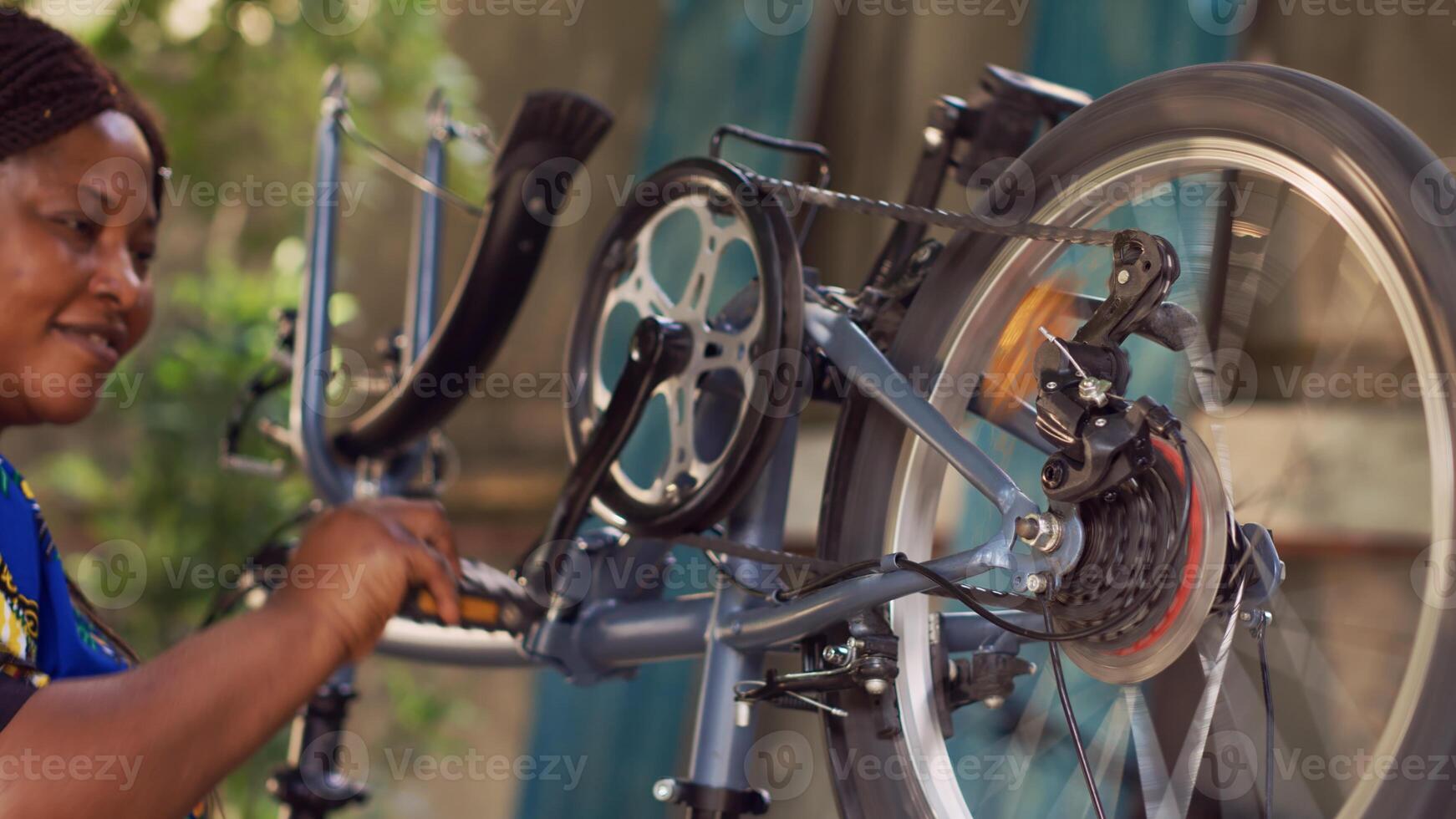 Young sports-loving black woman holding and adjusting bike components in home yard. Enthusiastic female cyclist thoroughly examining and fixing damaged bicycle for summer leisure cycling. photo