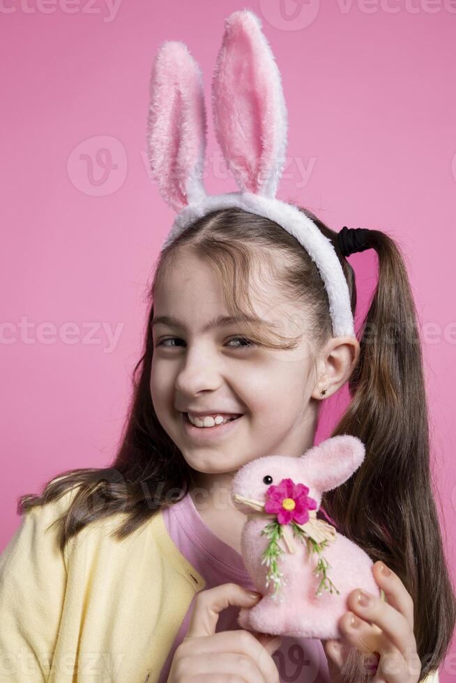 dulce pequeño niña posando con un linda rosado relleno Conejo en cámara, demostración su Pascua de Resurrección temática juguetes y decoraciones pequeño niño con conejito orejas sensación contento y emocionado acerca de celebracion. foto