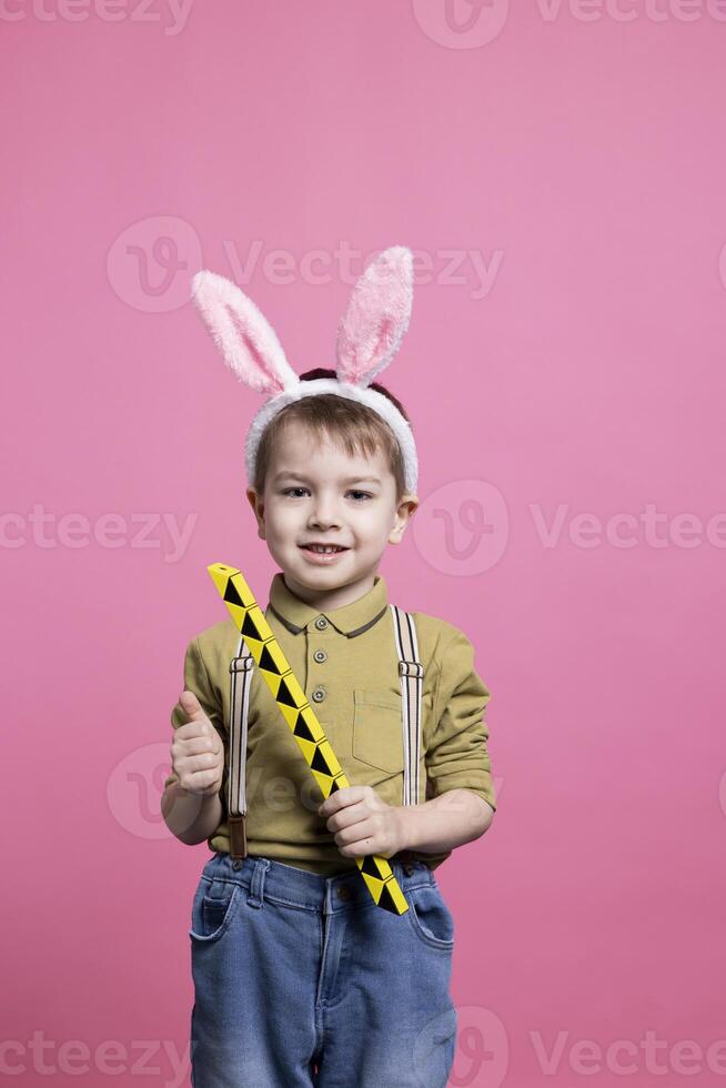 Young cute toddler playing around with a toy in front of camera, feeling happy while he wears fluffy bunny ears. Joyful small child posing against pink background, innocent kid. photo