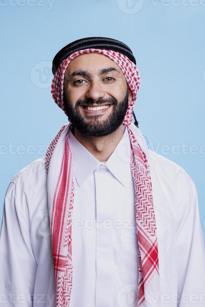 Cheerful muslim man dressed in islamic white thobe and checkered ghutra headdress studio portrait. Happy arab person standing and looking at camera with carefree expression photo