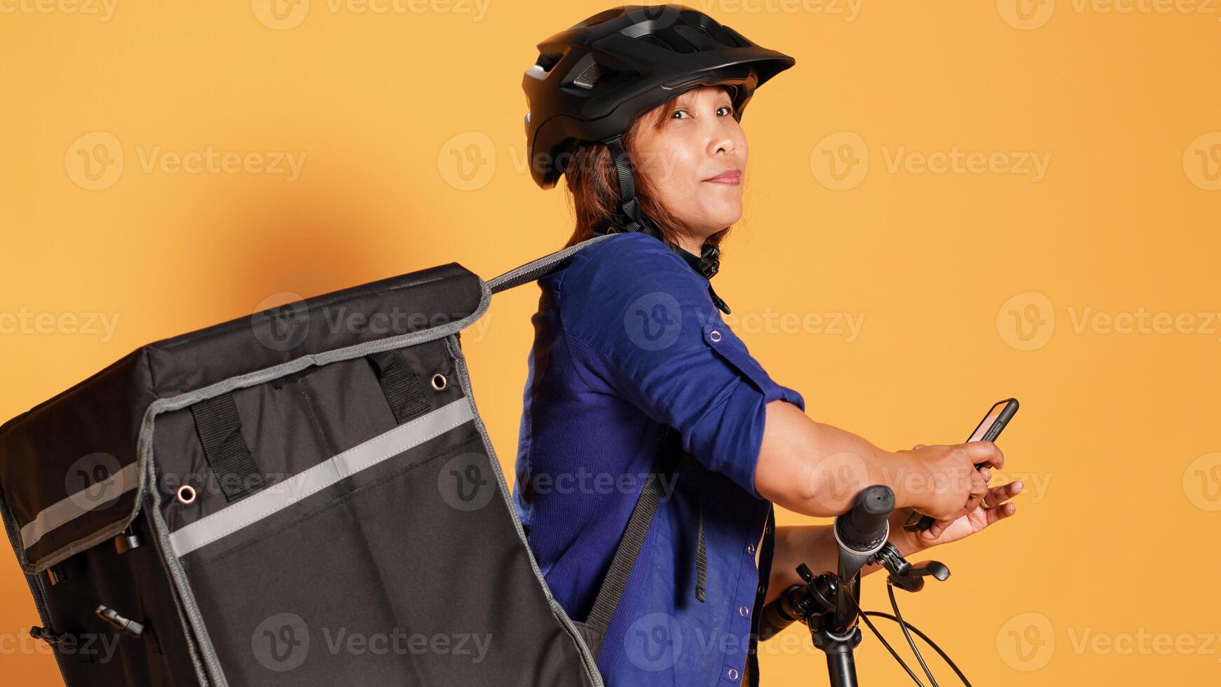 Tilt up panning camera shot of asian courier taking a break between food deliveries, texting friends. Bicycle rider waiting to receive next takeaway meal order, isolated over orange studio background photo
