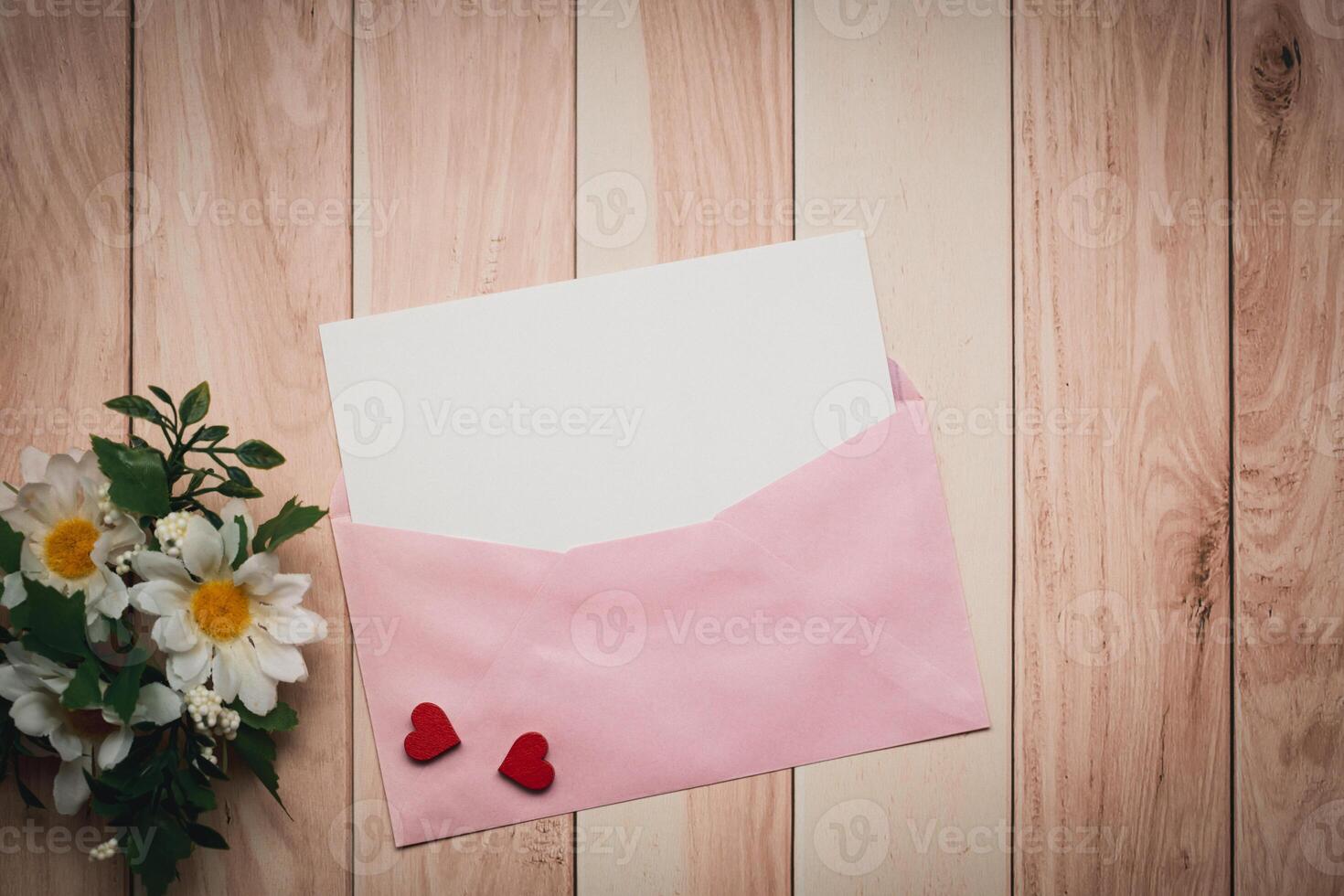 A pink envelope and a white card with a red heart and daisy are placed on a wooden table photo