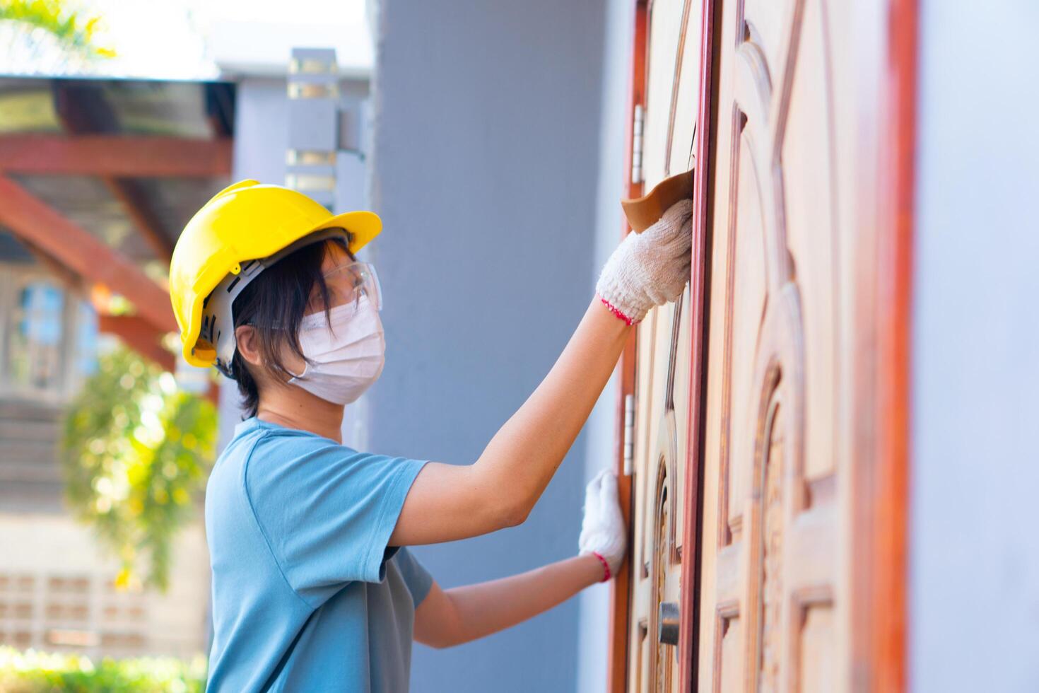 asiático hembra trabajador vistiendo un la seguridad casco utilizando papel de lija a polaco un de madera puerta a hogar. foto