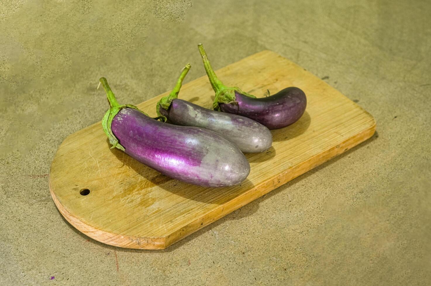 three purple eggplants or Solanum Melongena lying on a cutting board with a plain wooden board background photo