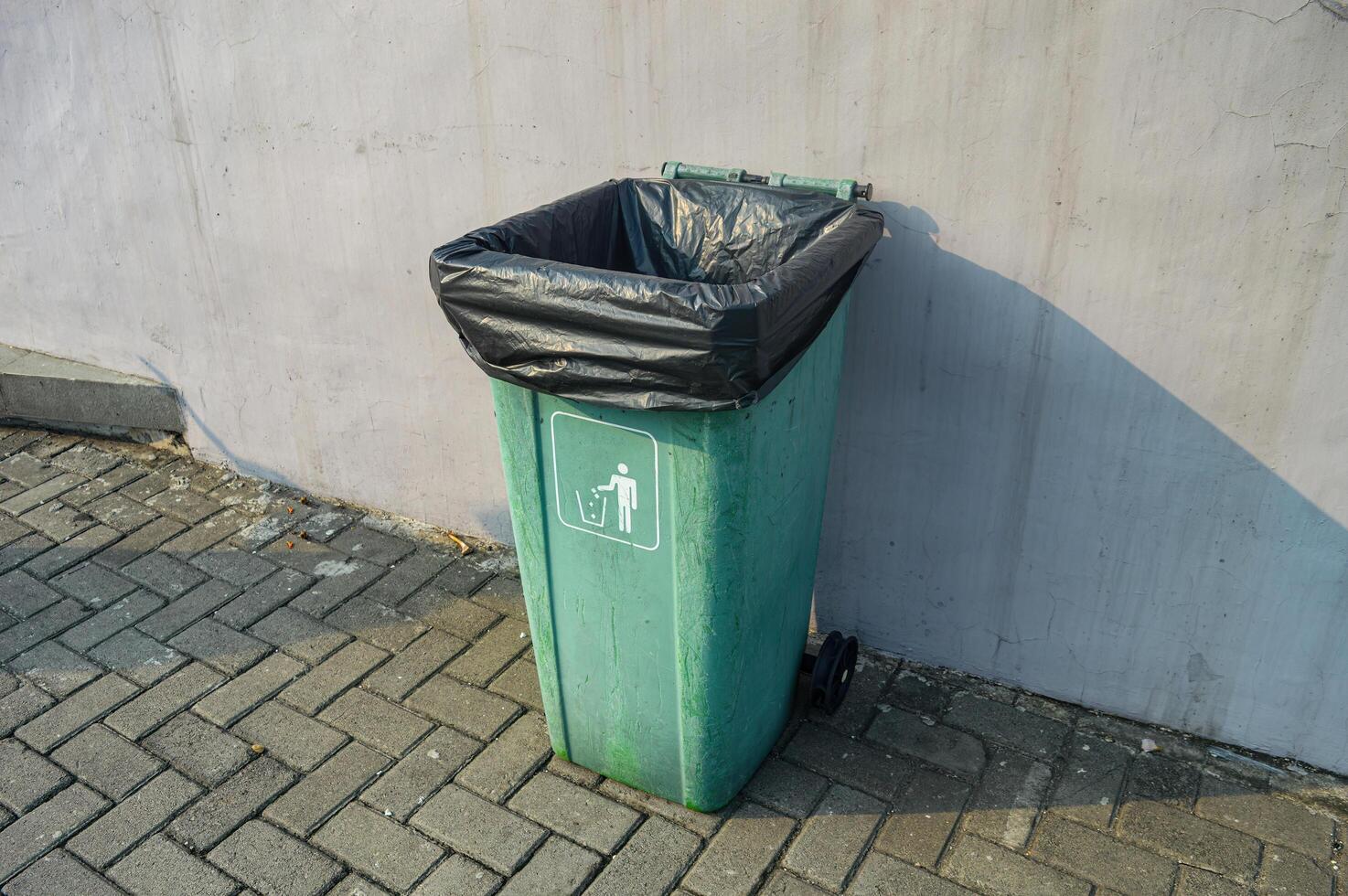 a green trash can with a black plastic trash bag inside on a paving block next to a white wall photo