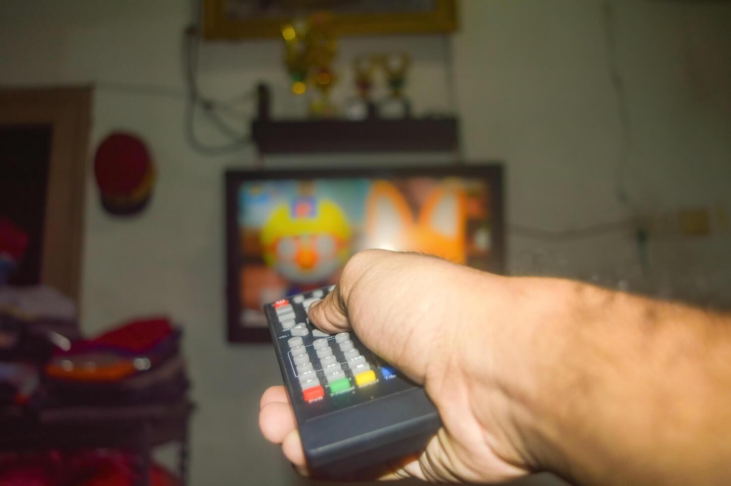 The hand is holding the TV remote in front of a television, preparing to change the TV channel photo