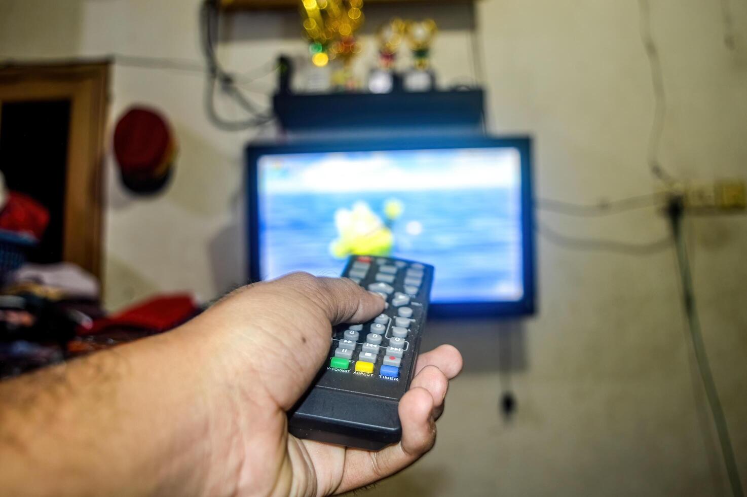 The hand is holding the TV remote in front of a television, preparing to change the TV channel photo