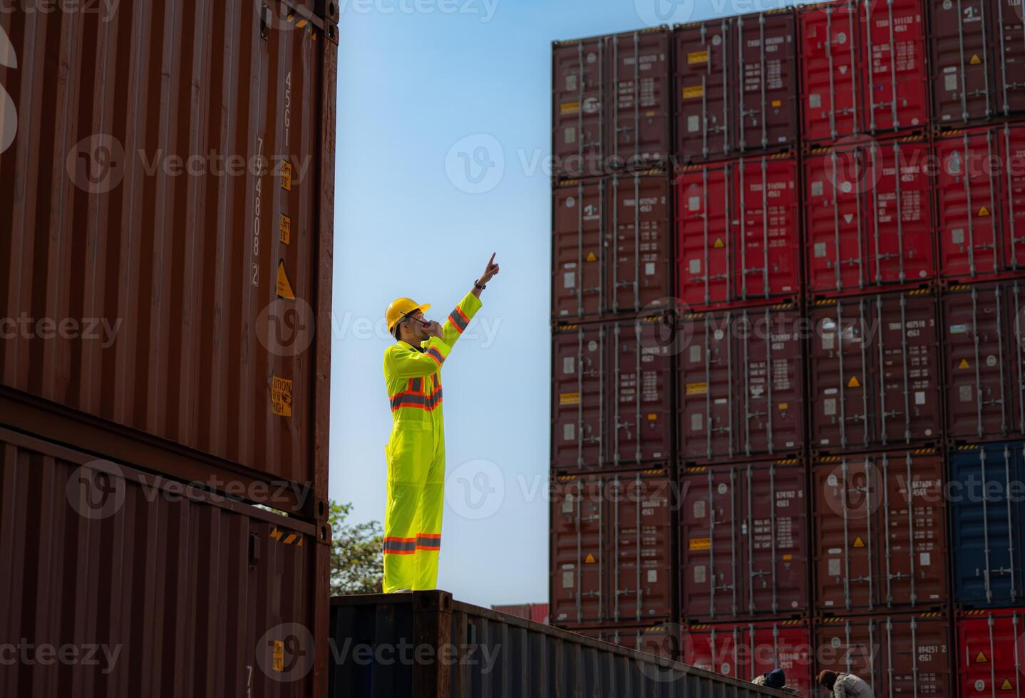 A male worker stands on a container box while wearing a safety suit. Use the walkie-talkie to communicate with coworkers on the floor below the container. photo