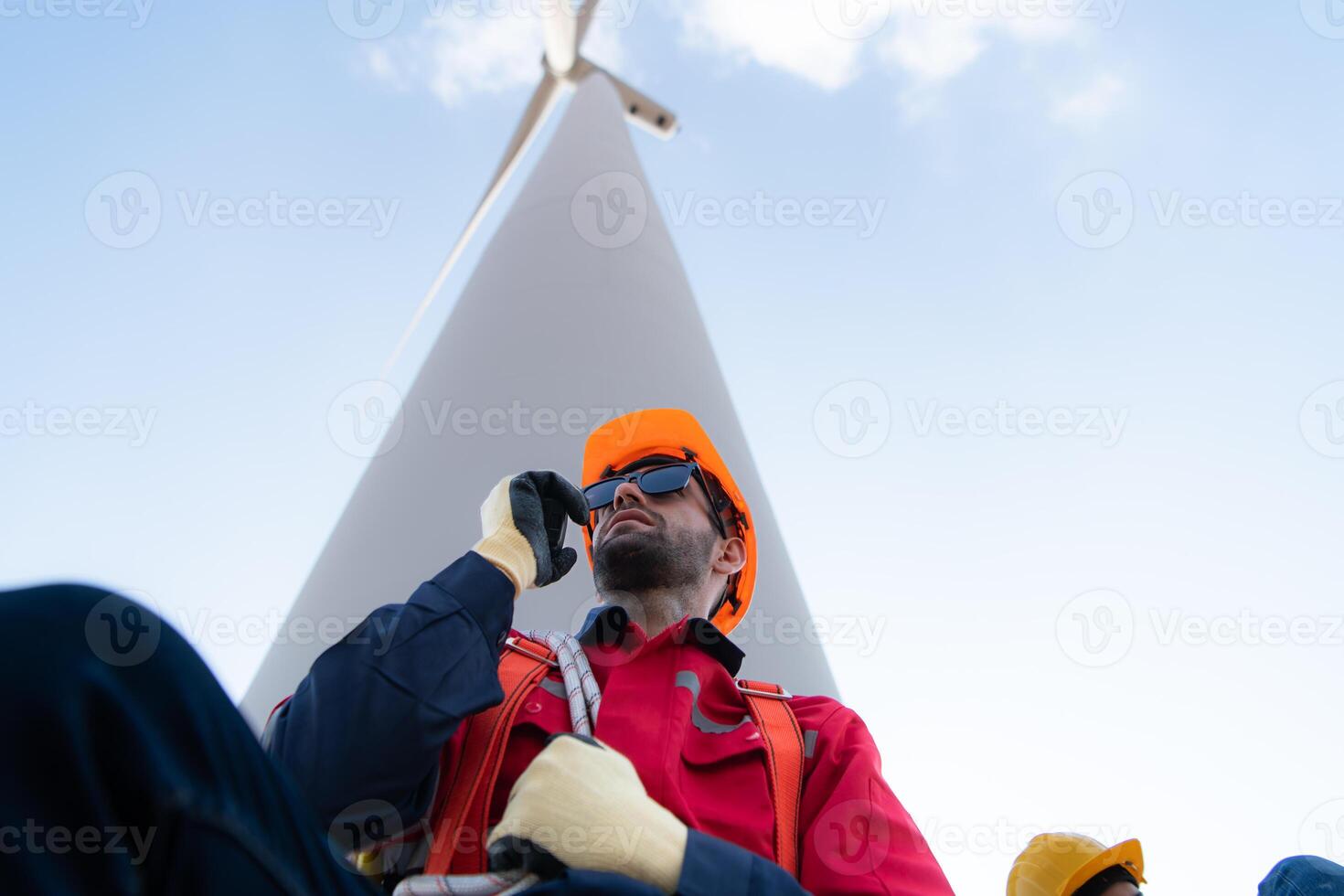 bajo ángulo ver de un masculino ingeniero en pie en frente de un viento turbina, utilizando el walkie película sonora a comunicar con compañeros de trabajo foto
