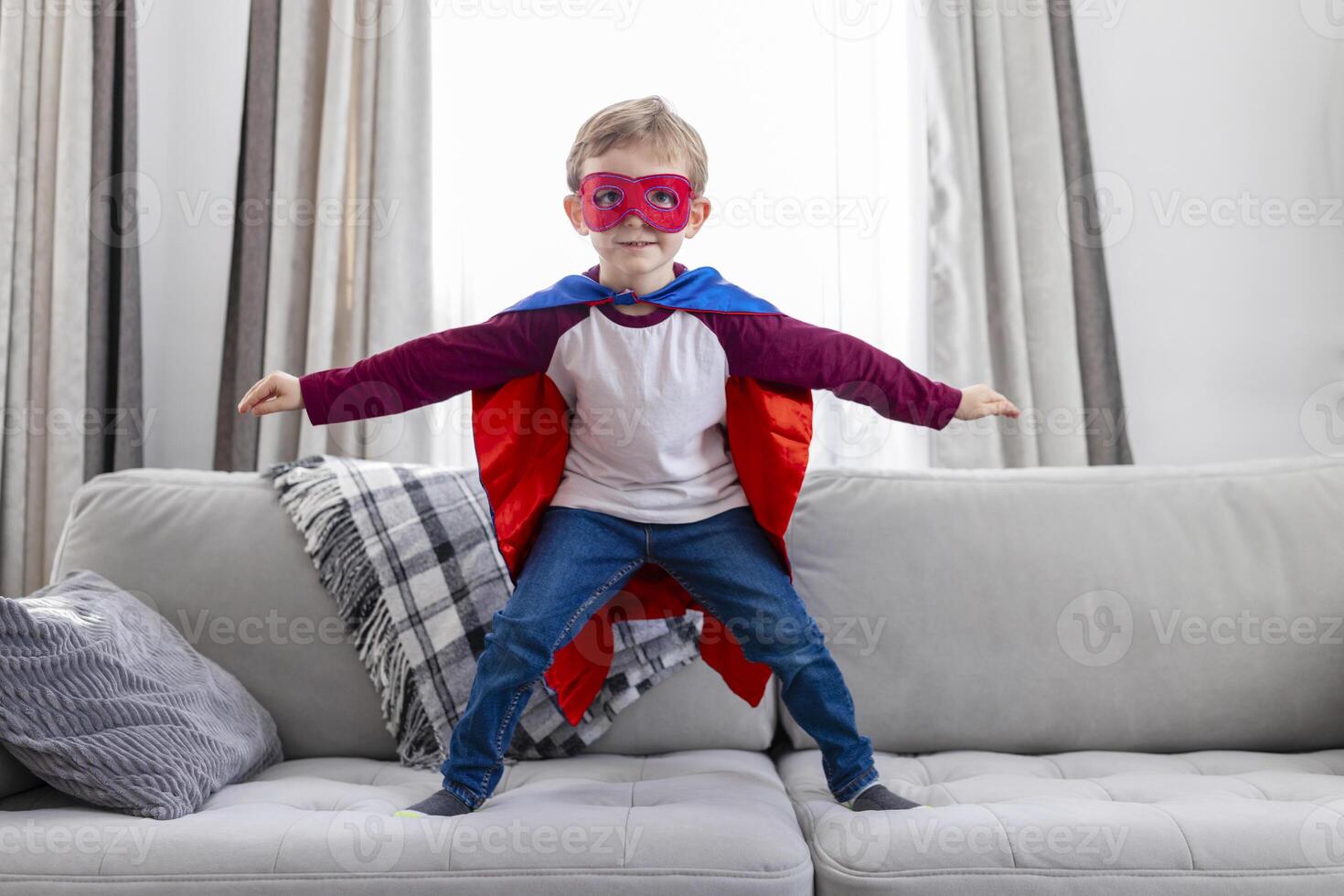 Boy in superhero costume with cape and mask standing on sofa photo