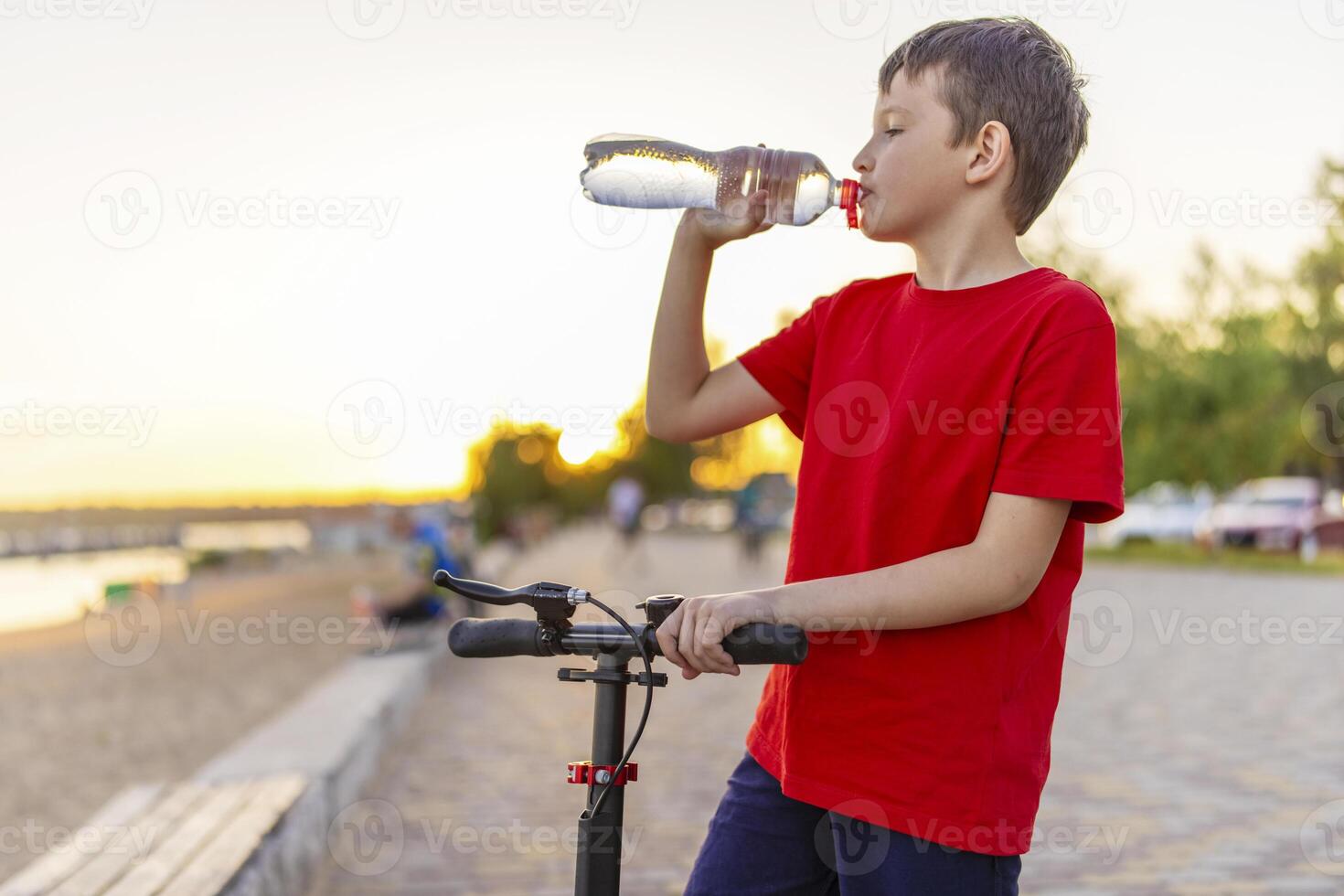 A boy drinks water from plastic bottle, standing with scooter photo