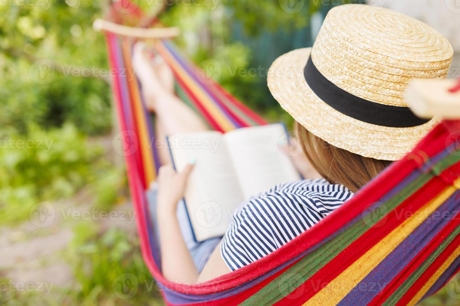 Young woman reading book while lying in comfortable hammock at green garden photo