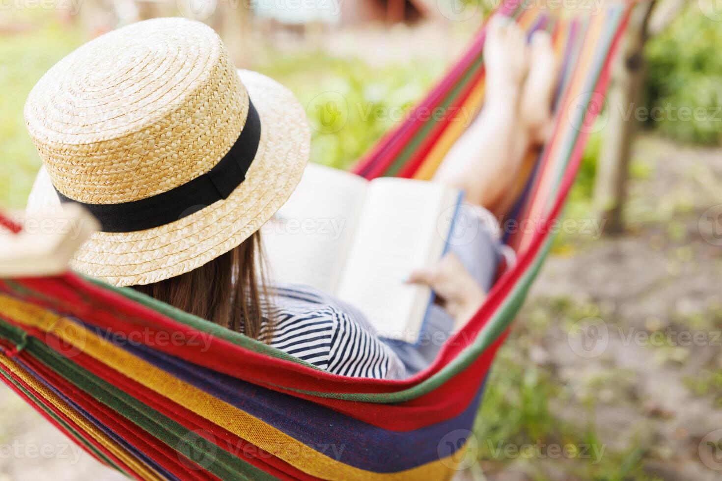 Young woman reading book while lying in comfortable hammock at green garden photo