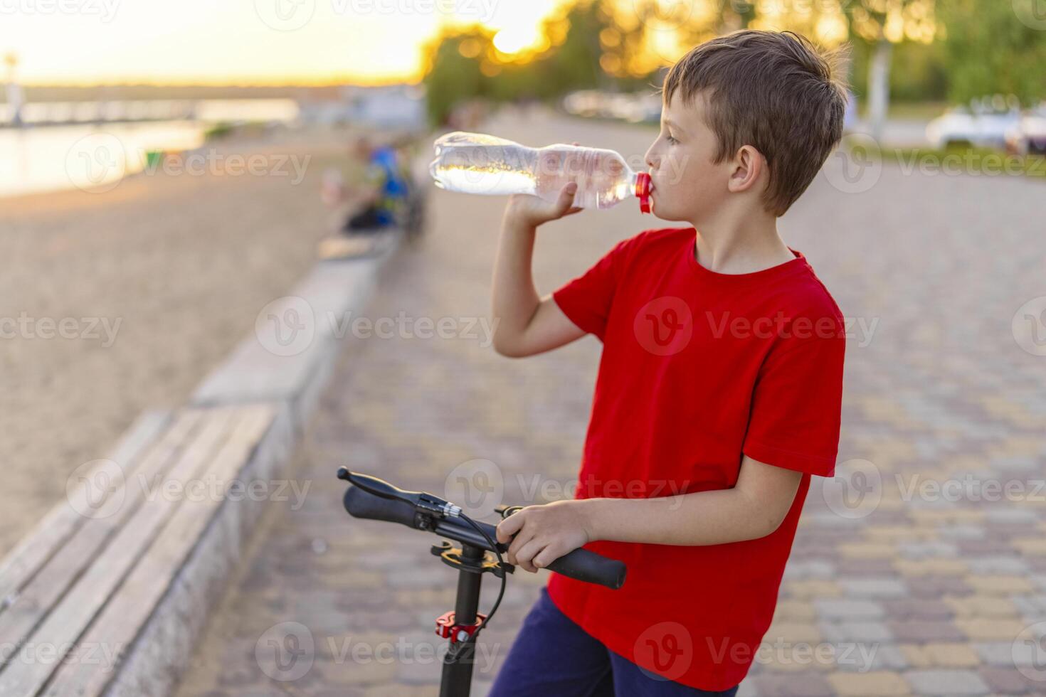 A boy drinks water from plastic bottle, standing with scooter photo